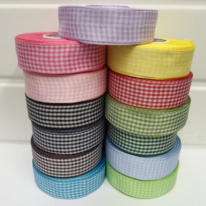 Op de afbeelding: Een stapel van twaalf rollen stoffen lint met een vichy-ruitpatroon in diverse kleuren, waaronder lavendel, roze, rood, geel, groen, blauw, zwart en bruin. Elke rol is cilindervormig met een zichtbare witte kartonnen kern in het midden.