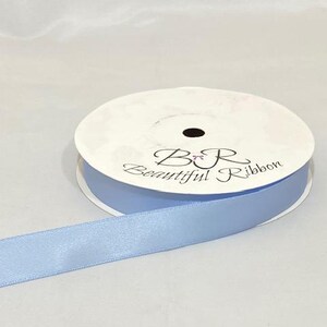 Puede incluir: Un carrete de cinta de raso azul claro con el texto "B&R Beautiful Ribbon" impreso en la parte superior. La cinta est&aacute; desenrollada, mostrando su textura suave y su color. El carrete es blanco con un agujero central negro.
