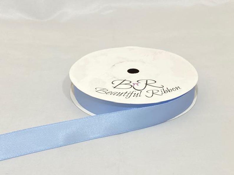 Puede incluir: Un carrete de cinta de raso azul claro con el texto "B&R Beautiful Ribbon" impreso en la parte superior. La cinta est&aacute; desenrollada, mostrando su textura suave y su color. El carrete es blanco con un agujero central negro.