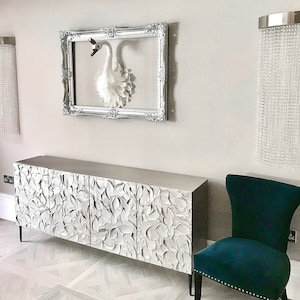 Puede incluir: Un aparador plateado, adornado y tallado con cuatro puertas. El aparador está en una habitación con un cisne blanco en un marco plateado en la pared y una silla azul turquesa.