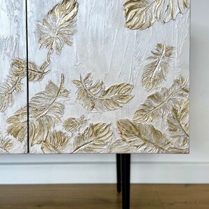 May include: A white cabinet with gold leaf feather details. The cabinet has two doors and sits on four black metal legs.