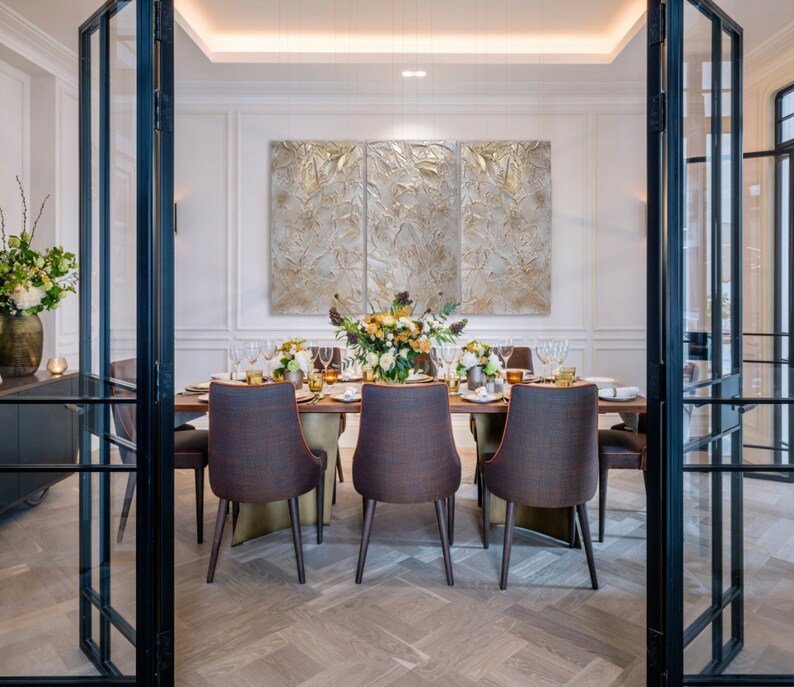 Textured triptych artwork in shades of  beige and gold, hangs on the wall of a dinengroom. Proudly highligting the wall above a dining table and chairs.