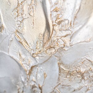Close up of a shimmering textured surface. The surface is rough, leafy like, painted in pearl white with gold contrast.