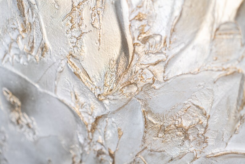 Close up of a shimmering textured surface. The surface is rough, leafy like, painted in pearl white with gold contrast.
