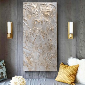 Textured abstract artwork in shades of silver and beige hangs on a gray wall between two wall sconces, above a bench with pillows and a small arrangement of white flowers.