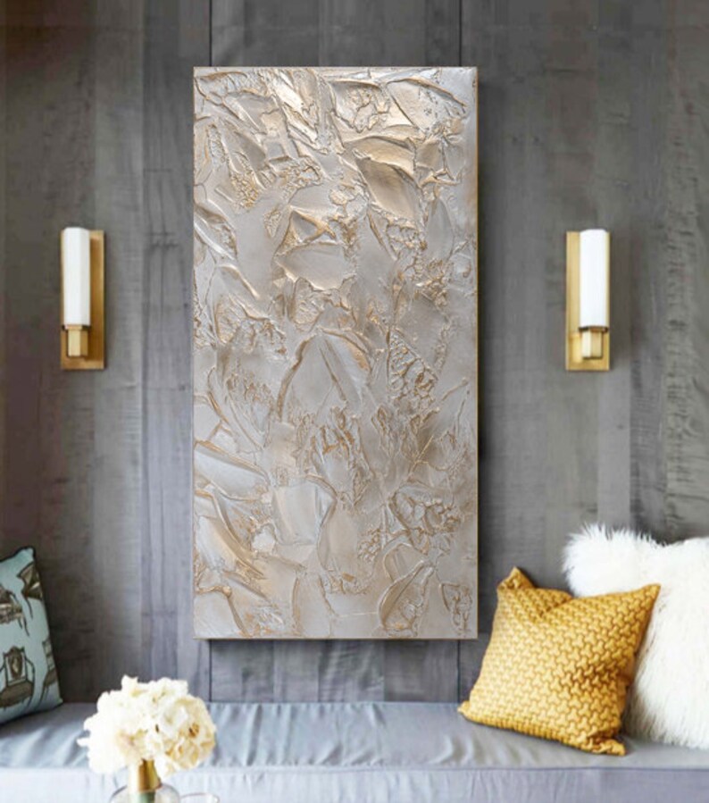 Textured abstract artwork in shades of silver and beige hangs on a gray wall between two wall sconces, above a bench with pillows and a small arrangement of white flowers.