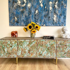 May include: A rustic, green and brown, four-door cabinet with gold metal legs. The cabinet is decorated with a marbled, distressed paint finish. The cabinet is sitting on a wood floor. There is a painting above the cabinet, a small elephant figurine, and a bust on top of the cabinet.