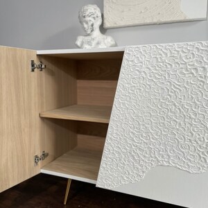 A custom-made sideboard with textured white doors, one of which is open to reveal wooden Oak interior with shelves inside. On top sits a white decorative bust alongside an abstract artwork on the wall, all elegantly set against a gray background.