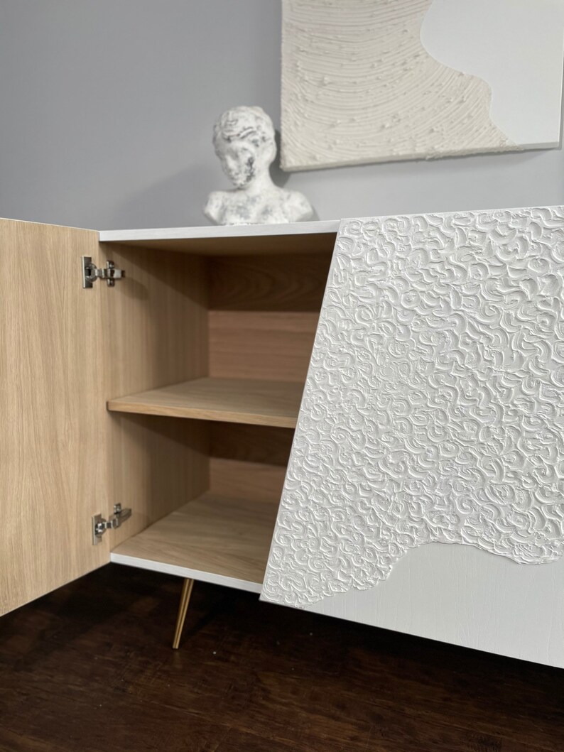 A custom-made sideboard with textured white doors, one of which is open to reveal wooden Oak interior with shelves inside. On top sits a white decorative bust alongside an abstract artwork on the wall, all elegantly set against a gray background.