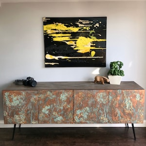 May include: A rustic wooden cabinet with three doors, each featuring a unique copper and turquoise textured design. The cabinet has black metal legs and sits on a wooden floor. A black and yellow abstract painting hangs on the wall above the cabinet.