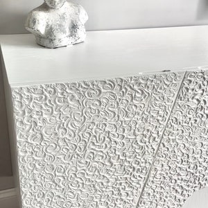 A custom-made sideboard with a decorative pattern stands on a wooden floor. On top, there's a white bust sculpture. A piece of art with a similar texture is partially visible above.