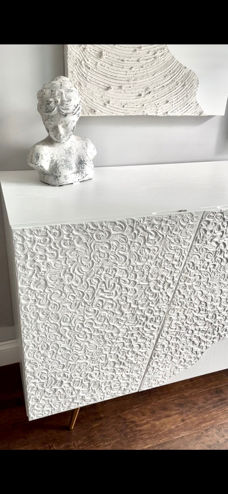A custom-made sideboard with a decorative pattern stands on a wooden floor. On top, there's a white bust sculpture. A piece of art with a similar texture is partially visible above.