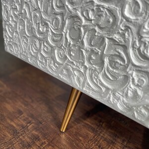 Close-up of a textured sideboard door finished in white.  A single slender brass leg is visible, supporting the bespoke furniture piece and adding a touch of elegance.