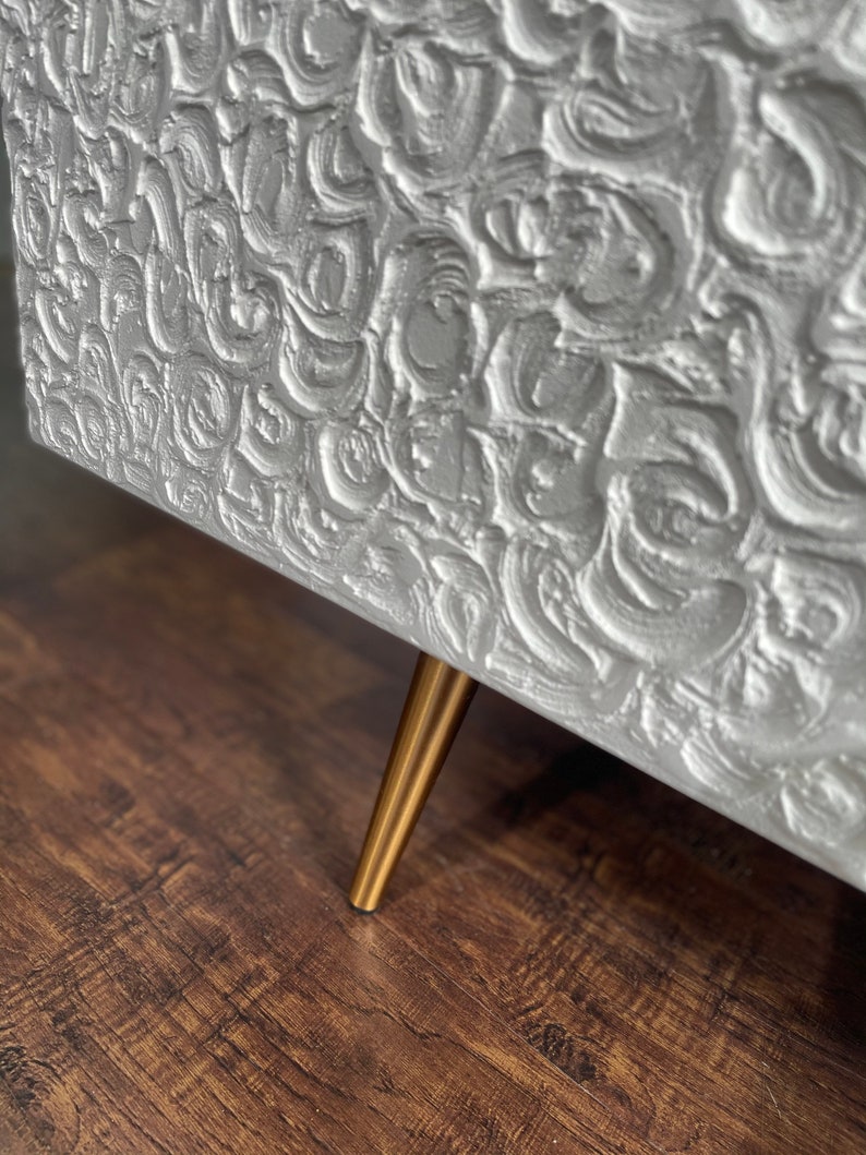 Close-up of a textured sideboard door finished in white.  A single slender brass leg is visible, supporting the bespoke furniture piece and adding a touch of elegance.