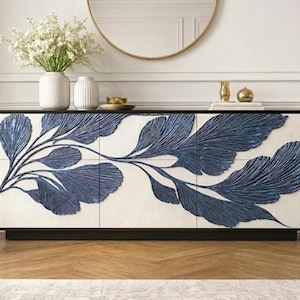 May include: A modern, low-profile cabinet with a black base and a white front panel. The panel features a large, stylized blue leaf design. The cabinet is topped with decorative items and a vase of white flowers. A gold-framed mirror hangs above.
