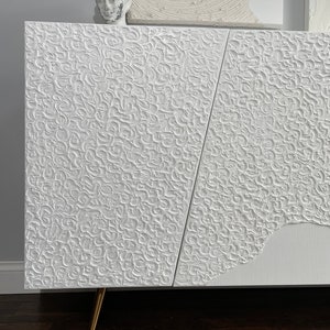 A bespoke white textured cabinet with abstract patterns stands on angled gold legs. A small sculpted head rests on top, while the background reveals a partial white textured artwork on the wall. This custom-made sideboard elegantly contrasts against the dark wood floor.