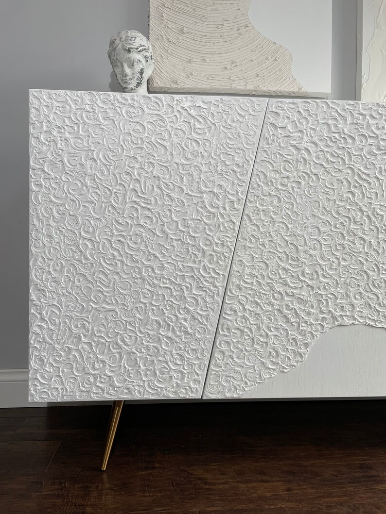 A bespoke white textured cabinet with abstract patterns stands on angled gold legs. A small sculpted head rests on top, while the background reveals a partial white textured artwork on the wall. This custom-made sideboard elegantly contrasts against the dark wood floor.