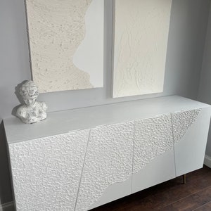 A modern room showcases bespoke furniture, with a white textured sideboard featuring an abstract pattern. Two textured abstract paintings hang on the grey wall above. A white sculpture bust is placed on the sideboard. The dark wood floor adds warmth to the space.