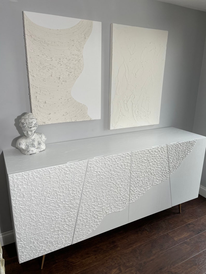 A modern room showcases bespoke furniture, with a white textured sideboard featuring an abstract pattern. Two textured abstract paintings hang on the grey wall above. A white sculpture bust is placed on the sideboard. The dark wood floor adds warmth to the space.