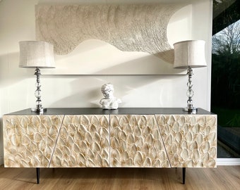May include: A white, textured, modern console table with three drawers. The table has a black top and black legs. Two lamps with white linen shades are on the table, and a white plaster sculpture is in the center.