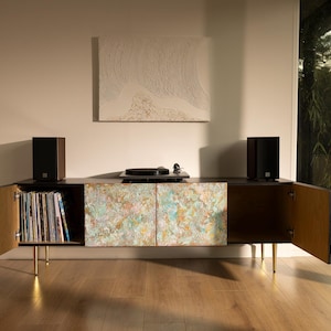 May include: A modern media console with a black top and sides, featuring doors with a colorful abstract design. The console has gold legs and open doors revealing vinyl records and a wooden interior. Two black speakers flank the console.