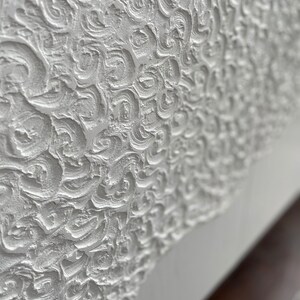 Close-up of a textured white surface with intricate swirls and patterns, reminiscent of bespoke furniture designs. The surface appears sculpted or embossed, creating a three-dimensional effect. At the bottom, theres a smooth transition to a plain white section.
