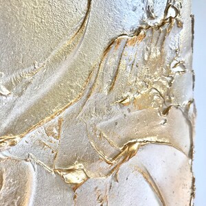 Close up of a shimmering textured surface. The surface is rough, leafy like, painted in pearl white with gold contrast.