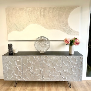 May include: A silver console table with four doors and a textured surface. The table has a black top and hairpin legs. The table is decorated with a silver decorative bowl and a vase of pink flowers.