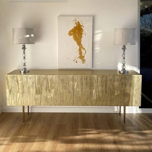 May include: A gold-colored, textured cabinet with four doors and thin, gold-colored legs. Two crystal-based lamps with gray shades sit on top of the cabinet. A white canvas with gold paint is mounted on the wall.