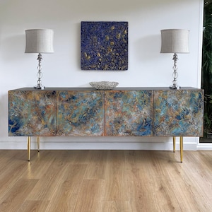 May include: A modern sideboard with a textured, abstract design in shades of blue, gold, and rust. The cabinet has four doors, gold legs, and is flanked by two lamps with gray shades. A blue and gold artwork hangs above the sideboard.