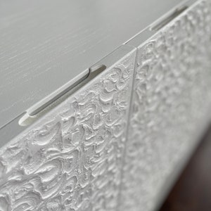 Close-up of a textured white cabinet showcasing intricate swirling patterns. This custom-made sideboard appears detailed and ornate, exuding elegance. Its smooth top edge allows light to reflect softly on the surface, enhancing its bespoke furniture appeal.
