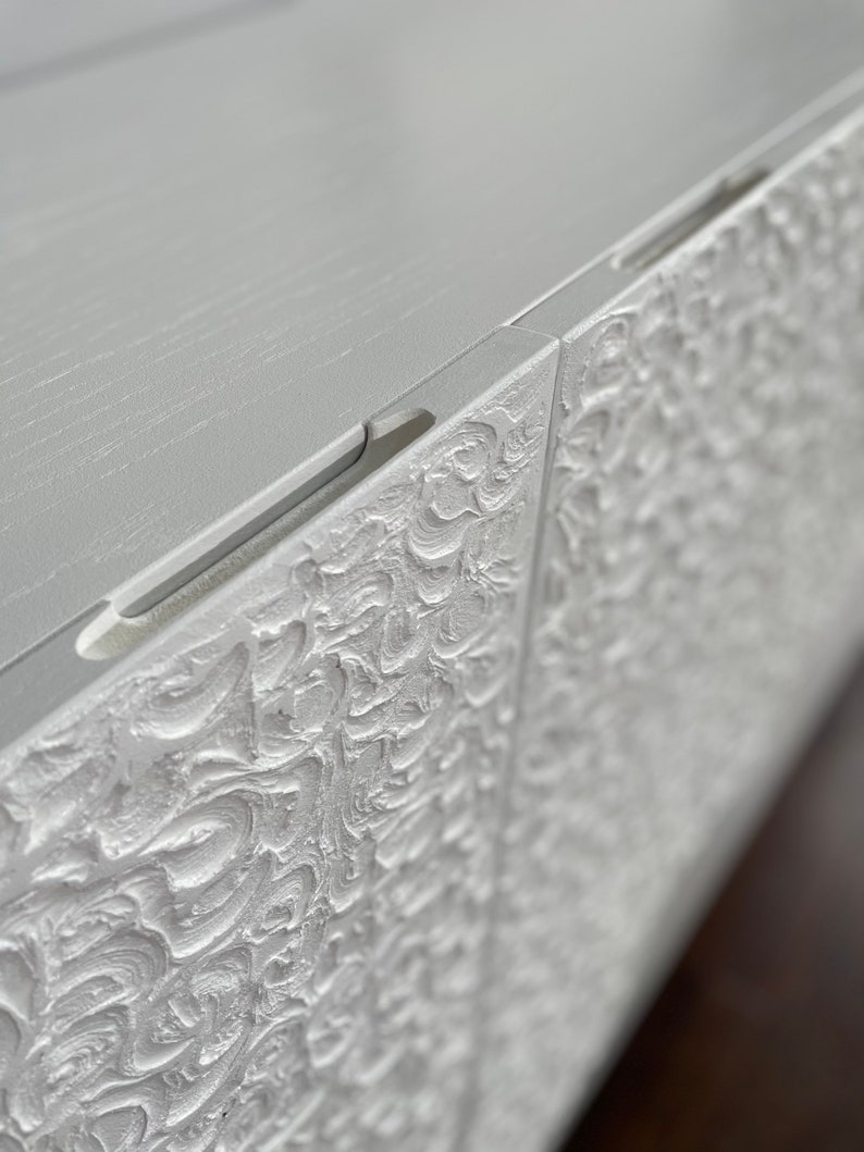 Close-up of a textured white cabinet showcasing intricate swirling patterns. This custom-made sideboard appears detailed and ornate, exuding elegance. Its smooth top edge allows light to reflect softly on the surface, enhancing its bespoke furniture appeal.