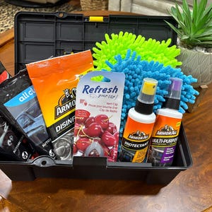 May include: A black plastic toolbox filled with car cleaning supplies. Includes Armor All protectant and multi-purpose cleaner, cherry-scented vent clips, microfiber wash mitts, and automotive cleaning products. A potted succulent sits on the side.