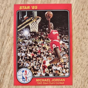 Puede incluir: Tarjeta de baloncesto roja con Michael Jordan en el concurso de mates de 1985. Incluye el texto "STAR '85" y el logo de la NBA. El fondo muestra una multitud de espectadores.