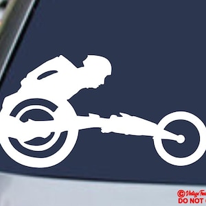 May include: White vinyl decal of a wheelchair racer in action, depicted in a dynamic, streamlined silhouette. The design features a racing wheelchair with large rear wheels and a smaller front wheel, set against a dark blue background.