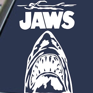 Op de afbeelding: Witte afbeelding van een open haaienbek met het woord "JAWS" erboven, tegen een donkerblauwe achtergrond. Het ontwerp bevat een zwemmer en een boot. De afbeelding is een sticker of decal.