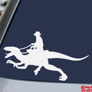 May include: White vinyl decal of a person riding a dinosaur, a humorous design. The rider wears a hat and holds reins. The dinosaur is a raptor with its mouth open. The decal is on a dark blue background.