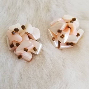 Pode incluir: Um par de brincos rosa e branco em forma de flor com detalhes marrons. Os brincos são feitos de plástico ou resina e têm um visual delicado e feminino.
