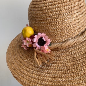 Pu&ograve; includere: Un cappello di paglia da sole con un'ampia tesa e una decorazione floreale e fruttata. Il cappello &egrave; di colore marrone chiaro e i fiori sono rosa e gialli.