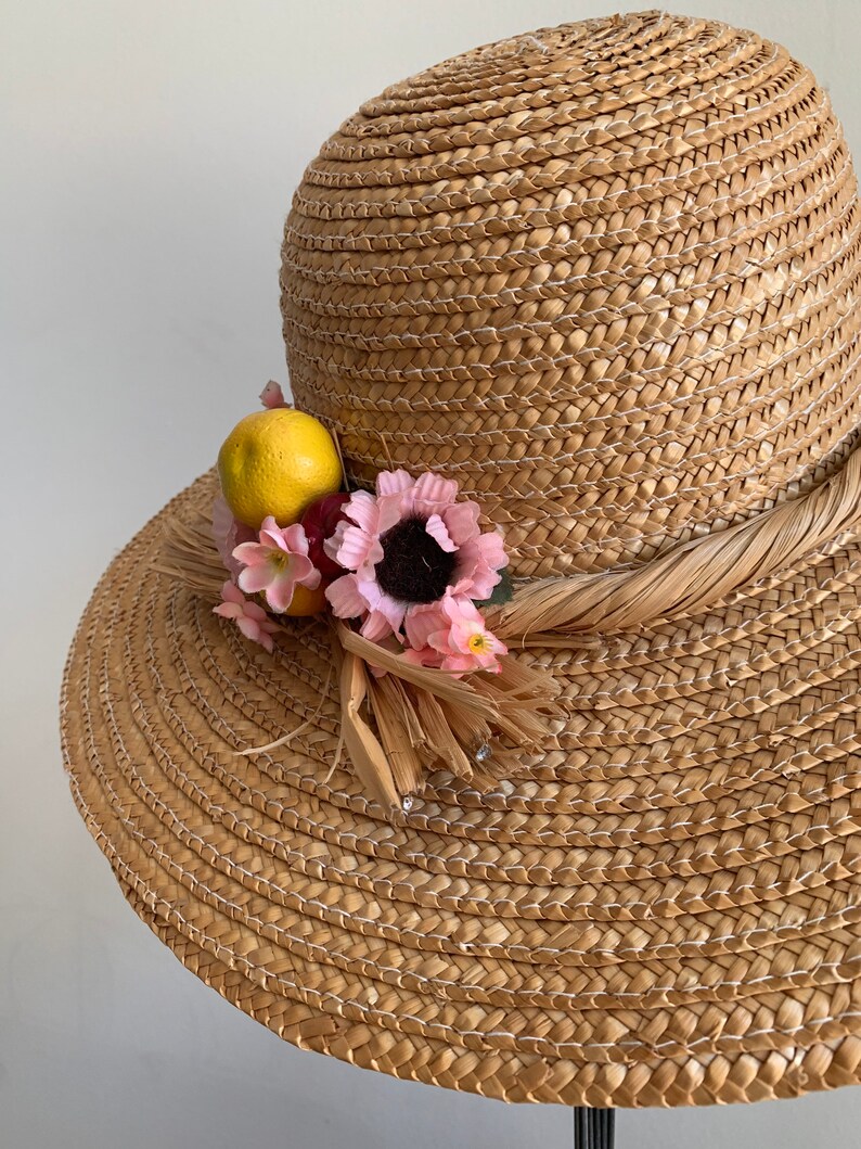 Pu&ograve; includere: Un cappello di paglia da sole con un'ampia tesa e una decorazione floreale e fruttata. Il cappello &egrave; di colore marrone chiaro e i fiori sono rosa e gialli.