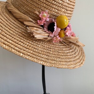 Pu&ograve; includere: Un cappello di paglia a tesa larga con una trama intrecciata marrone. Il cappello &egrave; decorato con fiori rosa, frutta gialla e arancione e dettagli in paglia marrone.