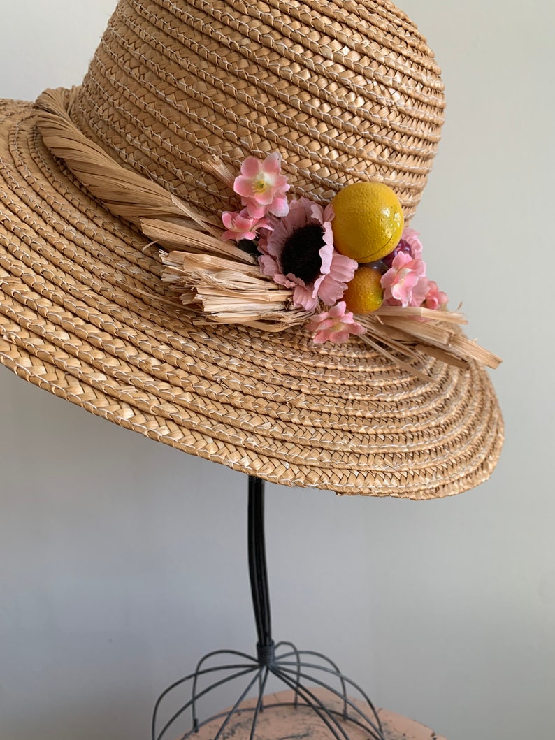Pu&ograve; includere: Un cappello di paglia a tesa larga con una trama intrecciata marrone. Il cappello &egrave; decorato con fiori rosa, frutta gialla e arancione e dettagli in paglia marrone.
