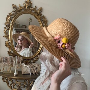 Pu&ograve; includere: Una donna che indossa un abito bianco con pizzo e un cappello di paglia con fiori rosa e limoni gialli. Il cappello ha un'ampia tesa ed &egrave; decorato con un nastro marrone.