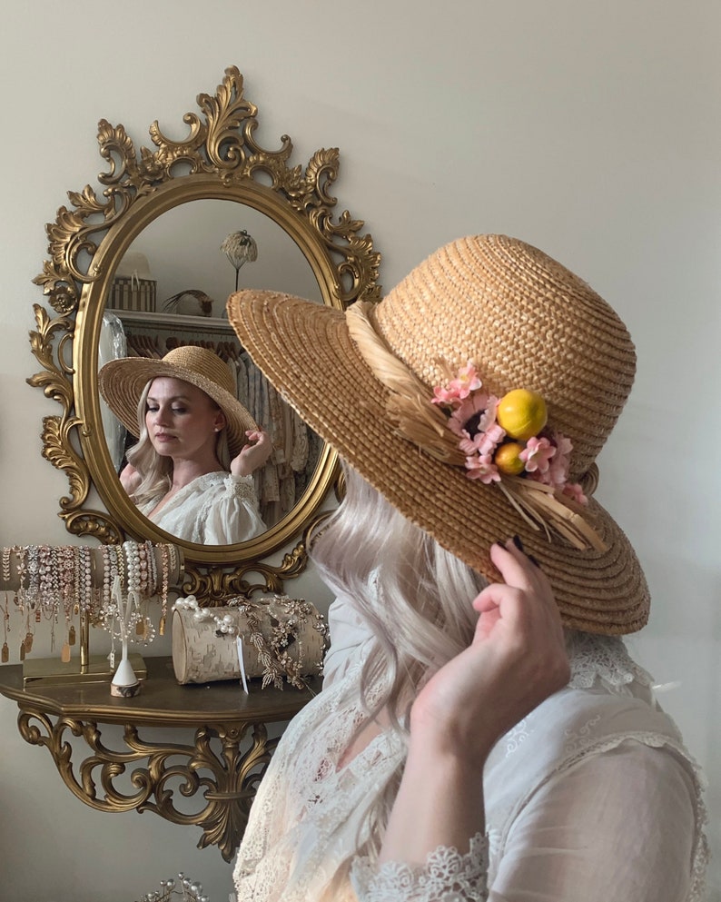 Pu&ograve; includere: Una donna che indossa un abito bianco con pizzo e un cappello di paglia con fiori rosa e limoni gialli. Il cappello ha un'ampia tesa ed &egrave; decorato con un nastro marrone.