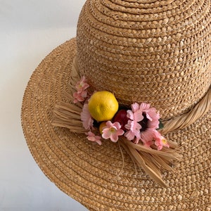 Pu&ograve; includere: Un cappello di paglia per il sole con un'ampia tesa e un bordo decorativo di fiori rosa e un limone giallo. Il cappello &egrave; fatto di paglia intrecciata e ha un colore marrone naturale.