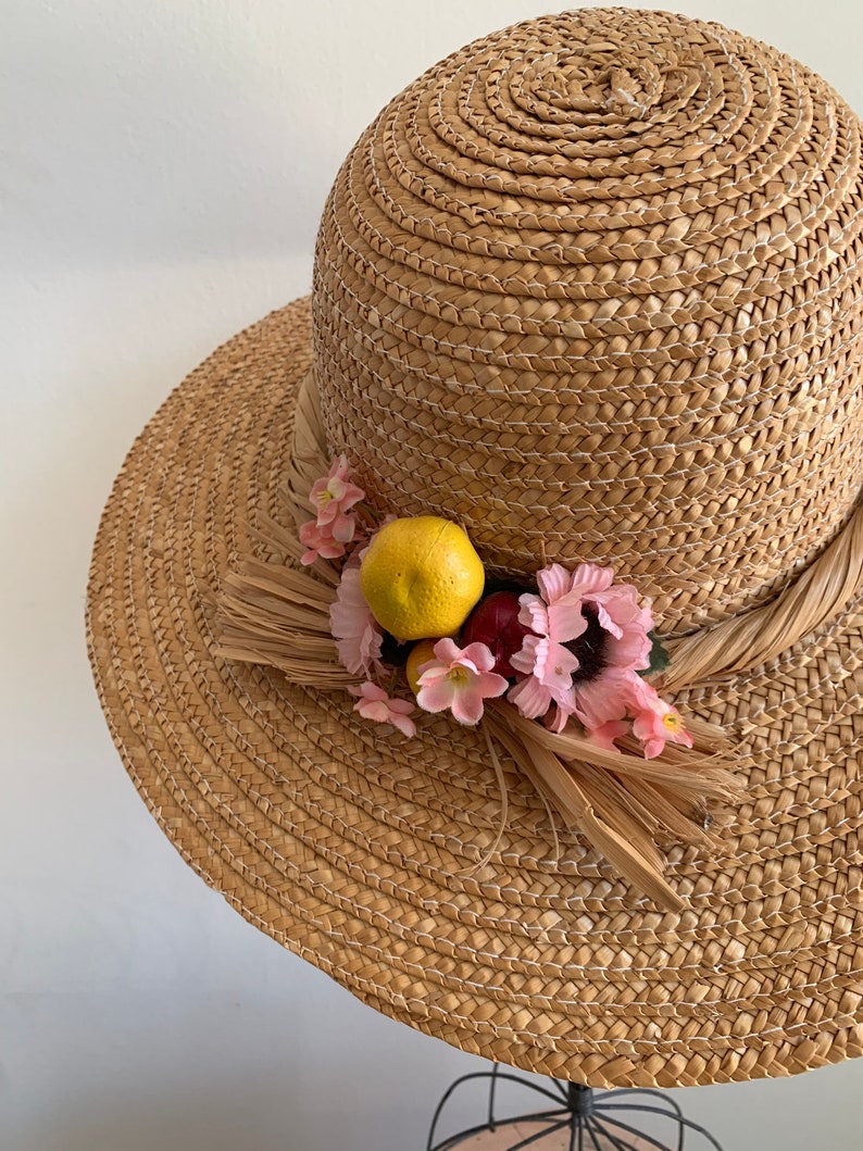 Pu&ograve; includere: Un cappello di paglia per il sole con un'ampia tesa e un bordo decorativo di fiori rosa e un limone giallo. Il cappello &egrave; fatto di paglia intrecciata e ha un colore marrone naturale.
