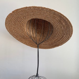 Pu&ograve; includere: Un cappello di paglia a tesa larga &egrave; esposto su un supporto per cappelli in filo di ferro nero. Il cappello &egrave; marrone e ha una trama intrecciata. Il supporto &egrave; fatto di filo sottile e ha una base rotonda.