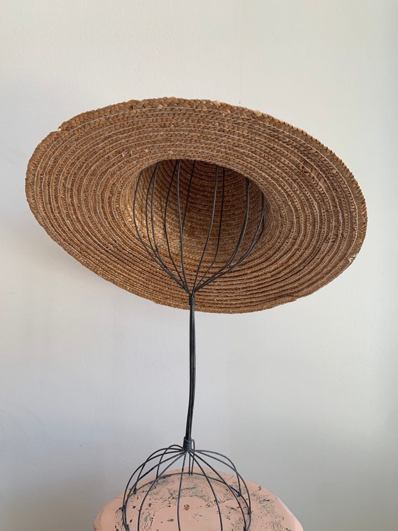 Pu&ograve; includere: Un cappello di paglia a tesa larga &egrave; esposto su un supporto per cappelli in filo di ferro nero. Il cappello &egrave; marrone e ha una trama intrecciata. Il supporto &egrave; fatto di filo sottile e ha una base rotonda.