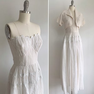 May include: A white strapless dress with a ruffled bodice and a layered skirt. The dress is paired with a matching white cropped jacket.