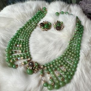 May include: A multi-strand green glass bead necklace with a gold tone clasp and matching earrings. The necklace has a large green glass bead with gold tone accents in the center.